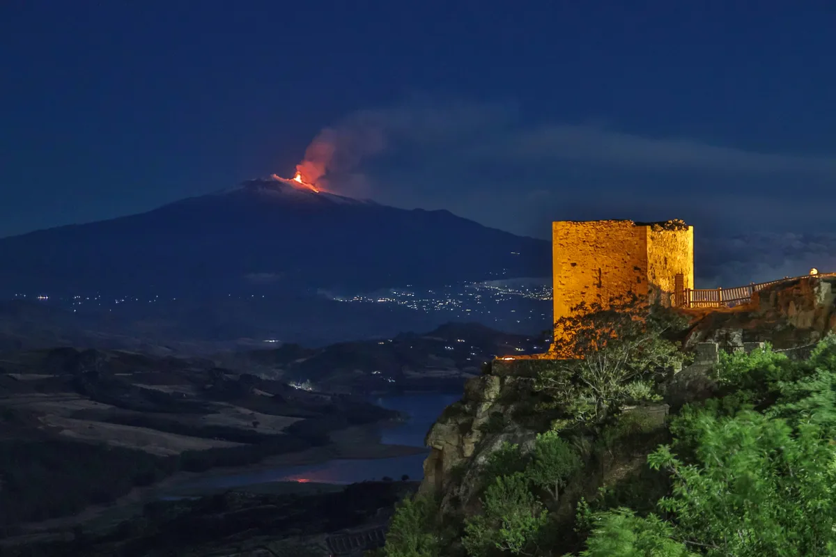 An illuminated tower of Agira Castle at night, with volcanic light on the horizon.